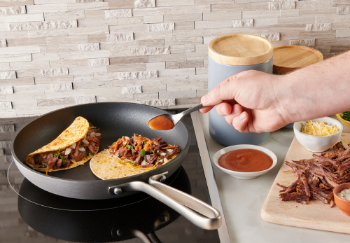 A person cooking in a pan, topping a tortilla with beef, cheese, onion, cilantro, and chipotle sauce, then folding it in half.